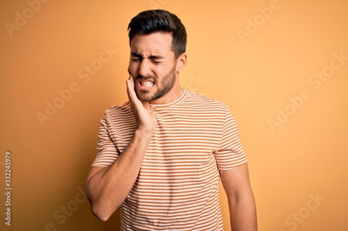Young handsome man with beard wearing casual striped t-shirt over yellow background touching mouth with hand with painful expression because of toothache or dental illness on teeth. Dentist photo