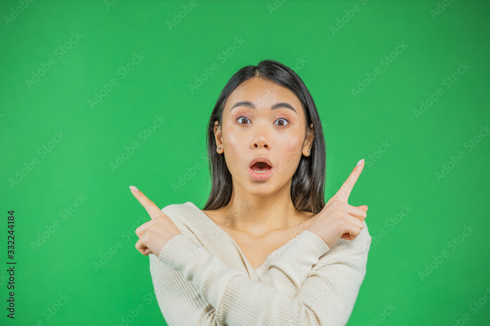 Smiling young asian woman in knitted shirt pointing over head and looking standing with thoughts before choosing. Waist up studio shot on green background Smiling young asian woman in knitted shirt pointing over head and looking standing with thoughts before choosing. Waist up studio shot on green background