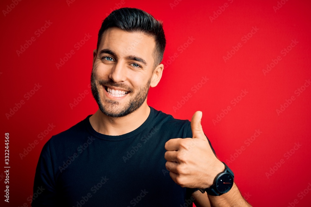 Young handsome man wearing casual black t-shirt standing over isolated red background doing happy thumbs up gesture with hand. Approving expression looking at the camera showing success. Young handsome man wearing casual black t-shirt standing over isolated red background doing happy thumbs up gesture with hand. Approving expression looking at the camera showing success.