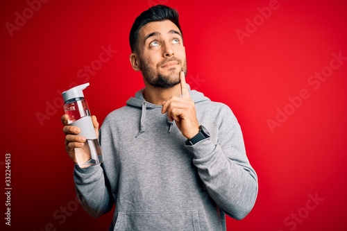 Young handsome sportsman drinking bottle of water to refeshment over red background serious face thinking about question, very confused idea © Krakenimages.com