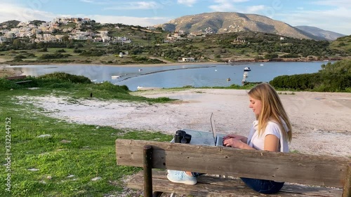 Young blond girl freelance remotely working on a laptop sitting on the bench with sea and mountain views
