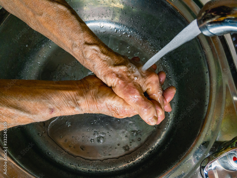  Hands of an elderly woman who washes her hands under a stream of water flowing from a tap.