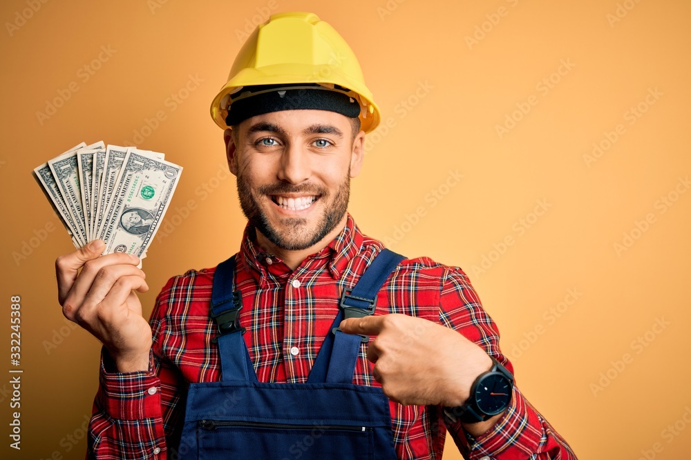 Young builder man wearing safety helmet holding dollars as payment over yellow background with surprise face pointing finger to himself Young builder man wearing safety helmet holding dollars as payment over yellow background with surprise face pointing finger to himself