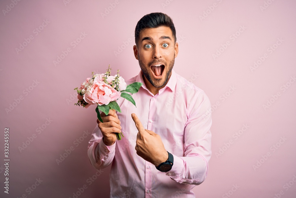 Young romatic man holding bouquet of spring flowers over pink isolatd background very happy pointing with hand and finger Young romatic man holding bouquet of spring flowers over pink isolatd background very happy pointing with hand and finger