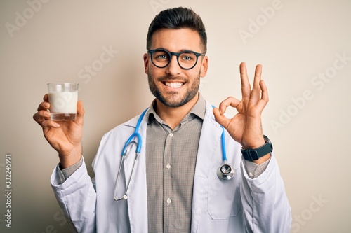 Young doctor man wearing stethoscope holding a glass of milk over isolated background doing ok sign with fingers, excellent symbol