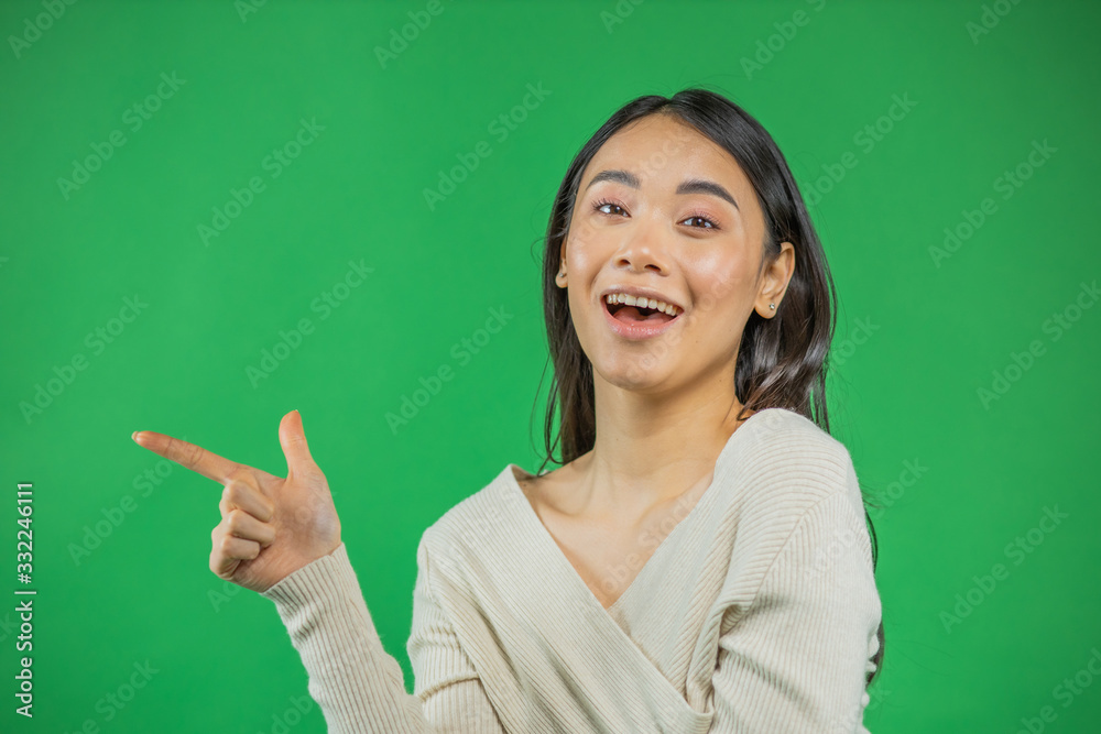 Smiling and confident woman in white is pointing to your product looking at the camera. Isolated on green background. Lovely mixed Asian, European model Smiling and confident woman in white is pointing to your product looking at the camera. Isolated on green background. Lovely mixed Asian, European model