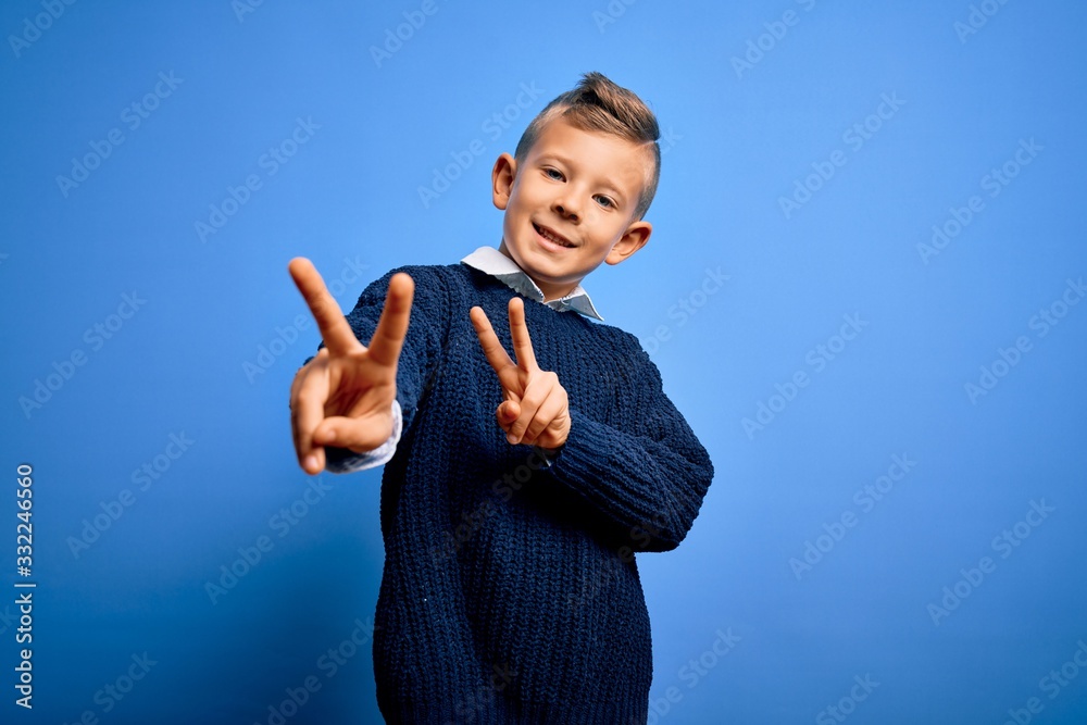 Young little caucasian kid with blue eyes wearing winter sweater over blue background smiling looking to the camera showing fingers doing victory sign. Number two. Young little caucasian kid with blue eyes wearing winter sweater over blue background smiling looking to the camera showing fingers doing victory sign. Number two.