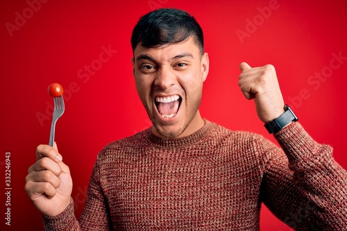 Young hispanic man holding fresh organic tomato on fork over red isolated background screaming proud and celebrating victory and success very excited, cheering emotion