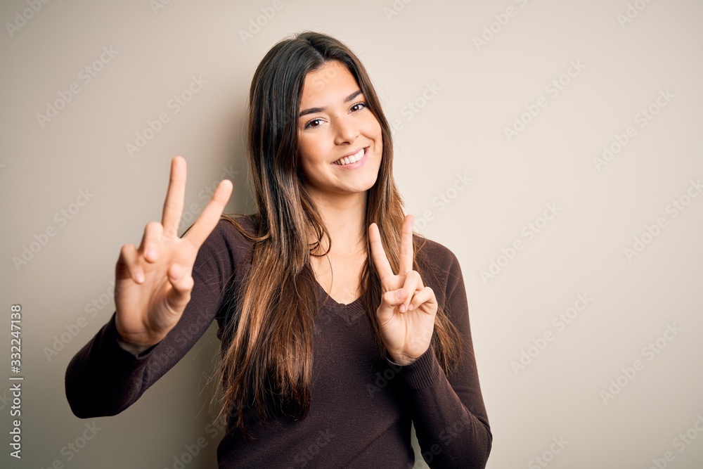 Young beautiful girl wearing casual sweater standing over isolated white background smiling looking to the camera showing fingers doing victory sign. Number two.