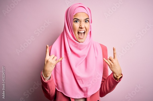 Young beautiful girl wearing muslim hijab standing over isolated pink background shouting with crazy expression doing rock symbol with hands up. Music star. Heavy music concept.