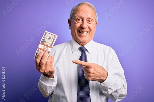 Senior grey haired man holding bunch of fifty dollars banknotes over purple background very happy pointing with hand and finger