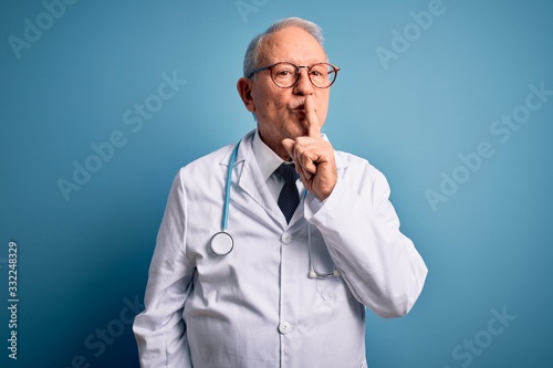 Senior grey haired doctor man wearing stethoscope and medical coat over blue background asking to be quiet with finger on lips. Silence and secret concept.