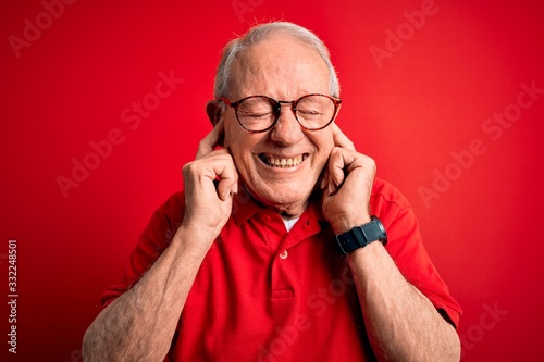 Grey haired senior man wearing glasses and casual t-shirt over red background covering ears with fingers with annoyed expression for the noise of loud music. Deaf concept.