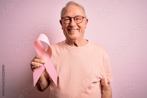 Grey haired senior man holding breast cancer awareness pink ribbon over pink background with a happy face standing and smiling with a confident smile showing teeth