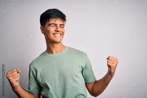 Young handsome man wearing casual t-shirt standing over isolated white background very happy and excited doing winner gesture with arms raised, smiling and screaming for success. Celebration concept.