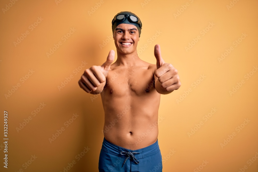 Young handsome man shirtless wearing swimsuit and swim cap over isolated yellow background approving doing positive gesture with hand, thumbs up smiling and happy for success. Winner gesture.
