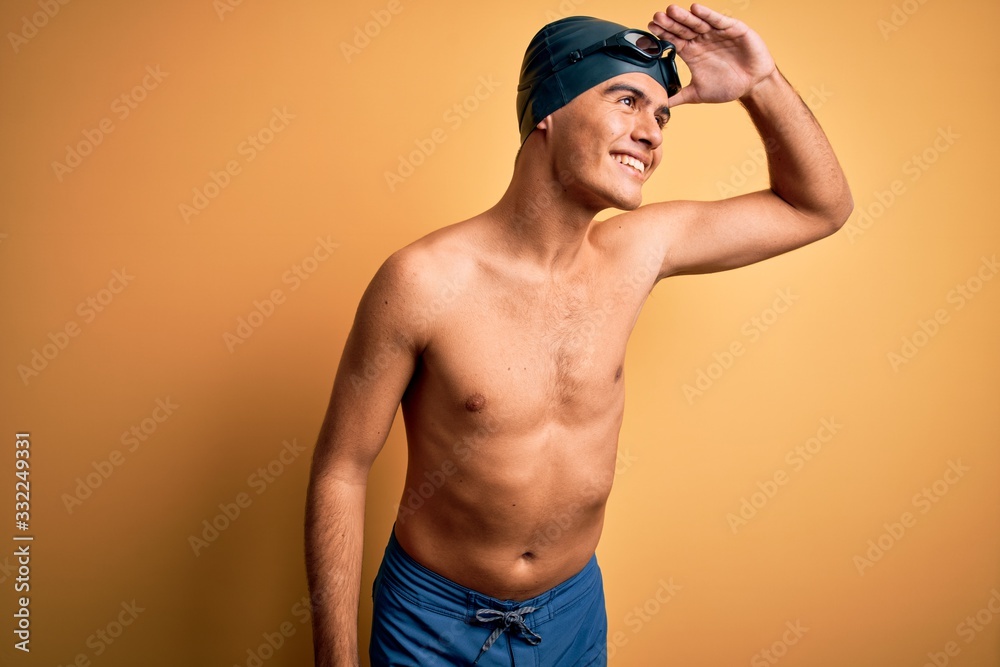 Young handsome man shirtless wearing swimsuit and swim cap over isolated yellow background very happy and smiling looking far away with hand over head. Searching concept.