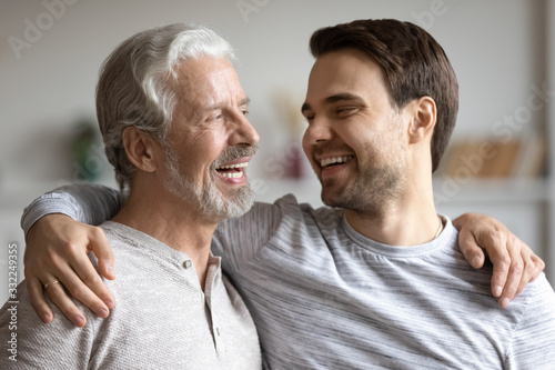 Overjoyed young millennial man embrace mature dad have fun talk and laugh at home, happy elderly father hug grown-up adult son enjoy family weekend reunion, show family love and care