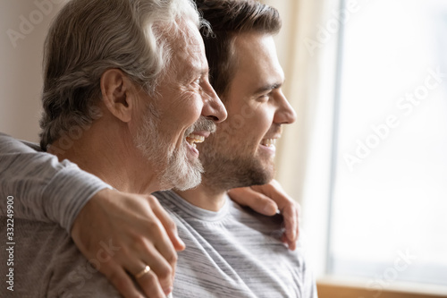 Close up of elderly father and adult grown-up son hug look in distance planning or dreaming together, happy hopeful young millennial man embrace mature dad thinking visualizing good future
