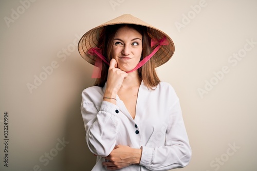 Young beautiful redhead woman wearing asian traditional conical hat over white background with hand on chin thinking about question, pensive expression. Smiling with thoughtful face. Doubt concept.