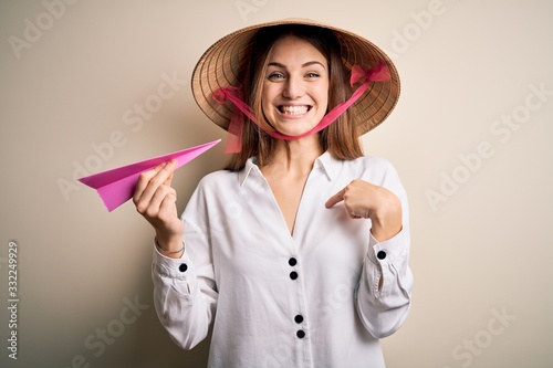 Young beautiful redhead woman wearing asian traditional hat holding paper airplane with surprise face pointing finger to himself