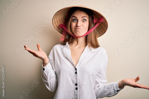 Young beautiful redhead woman wearing asian traditional conical hat over white background clueless and confused expression with arms and hands raised. Doubt concept.