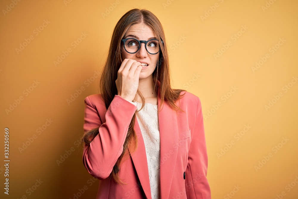 Young beautiful redhead woman wearing jacket and glasses over isolated yellow background looking stressed and nervous with hands on mouth biting nails. Anxiety problem. Young beautiful redhead woman wearing jacket and glasses over isolated yellow background looking stressed and nervous with hands on mouth biting nails. Anxiety problem.