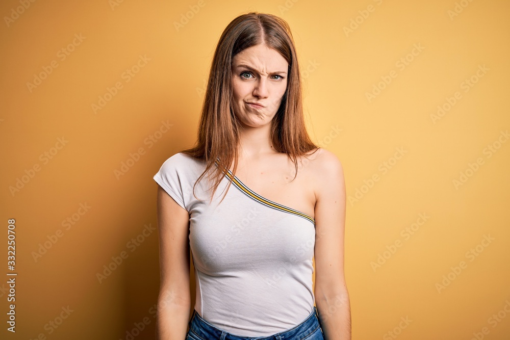 Young beautiful redhead woman wearing casual t-shirt over isolated yellow background skeptic and nervous, frowning upset because of problem. Negative person. Young beautiful redhead woman wearing casual t-shirt over isolated yellow background skeptic and nervous, frowning upset because of problem. Negative person.