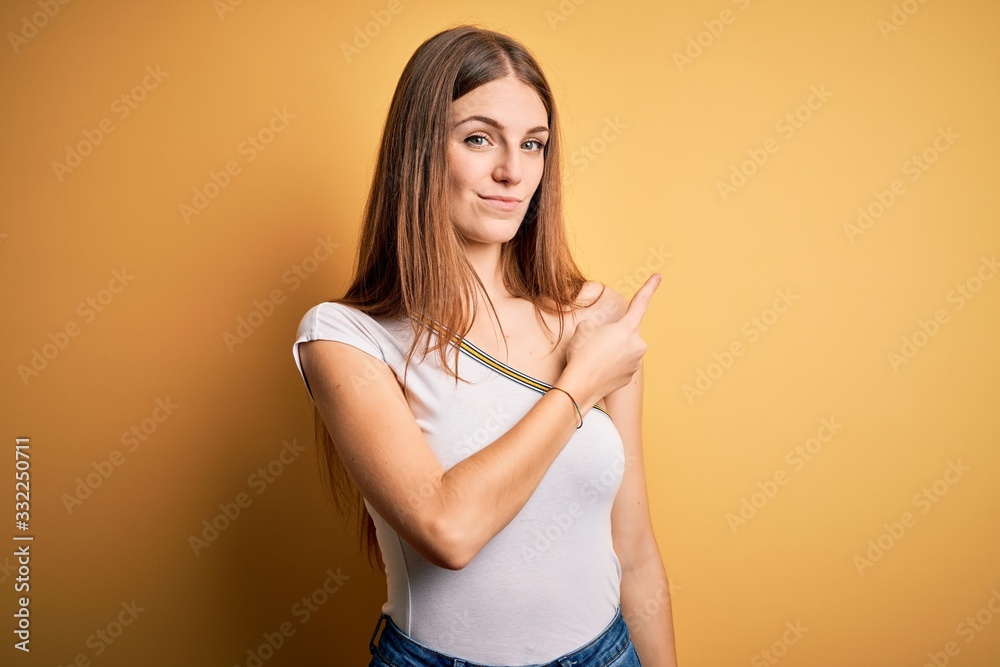 Young beautiful redhead woman wearing casual t-shirt over isolated yellow background Pointing with hand finger to the side showing advertisement, serious and calm face Young beautiful redhead woman wearing casual t-shirt over isolated yellow background Pointing with hand finger to the side showing advertisement, serious and calm face