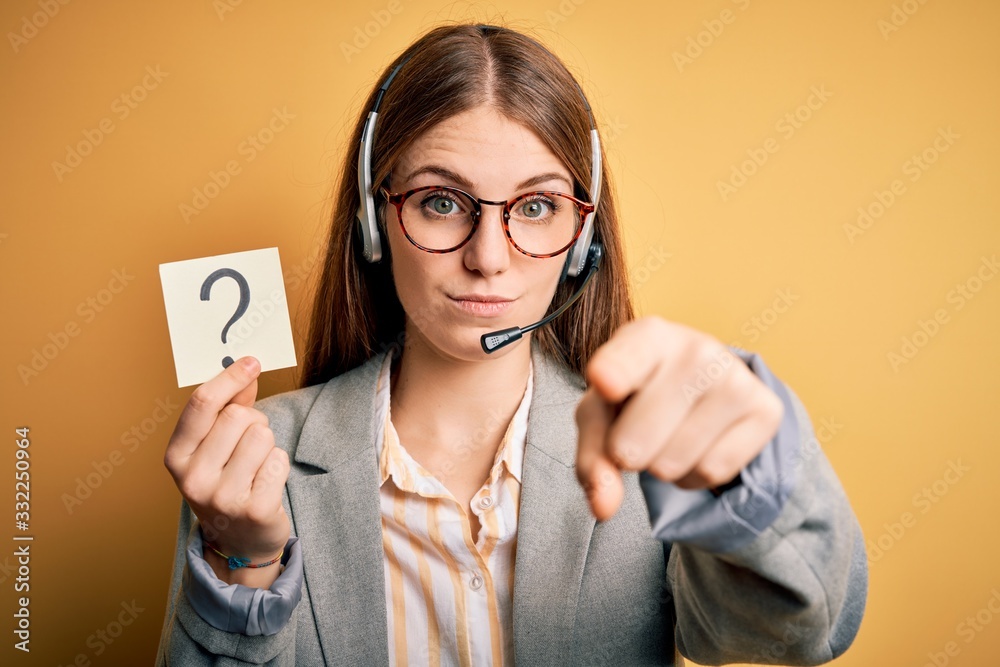 Young redhead call center agent woman using headset holding question mark reminder pointing with finger to the camera and to you, hand sign, positive and confident gesture from the front Young redhead call center agent woman using headset holding question mark reminder pointing with finger to the camera and to you, hand sign, positive and confident gesture from the front
