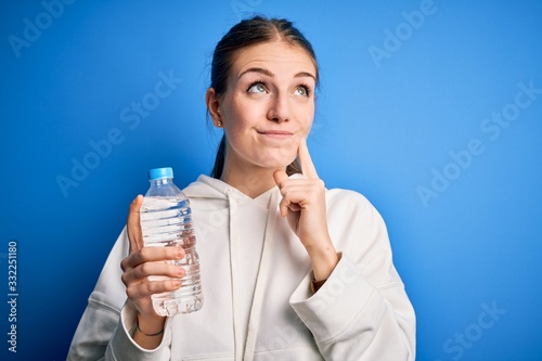 Young beautiful redhead woman doing sport drinking bottle of water over blue background serious face thinking about question, very confused idea photo