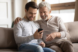 Overjoyed elderly father relax on sofa with adult son laugh watch funny videos on cellphone together, happy mature dad rest on couch at home with grown-up man child have fun using modern smartphone