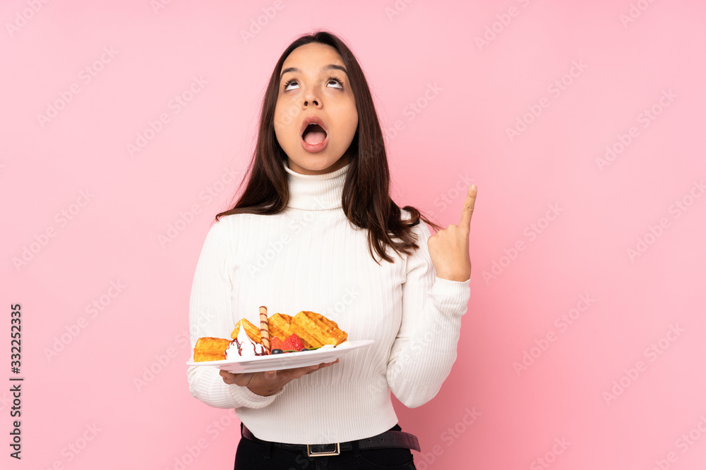 Young brunette woman holding waffles over isolated pink background pointing up and surprised