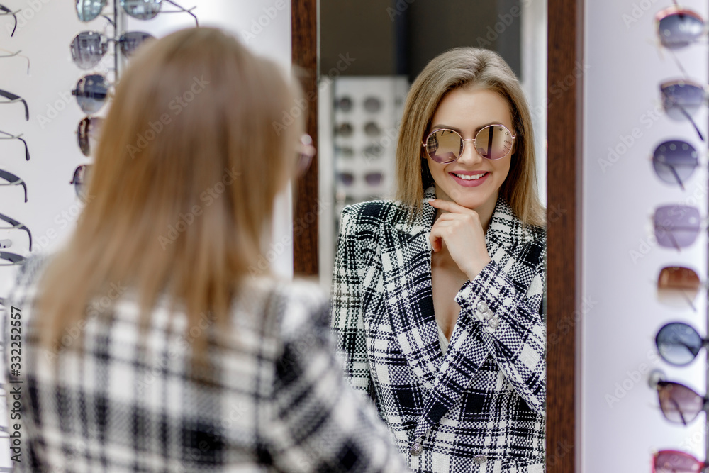 Beautiful and preety woman choosing a glasses in optician store, female looking at the mirror and smiling Beautiful and preety woman choosing a glasses in optician store, female looking at the mirror and smiling