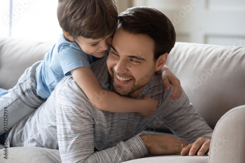 Cute little boy lying on happy young father hugging relaxing together on sofa in living room, overjoyed small preschooler son embrace loving dad, show affection, enjoy family weekend at home