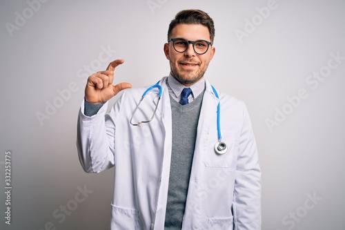 Young doctor man with blue eyes wearing medical coat and stethoscope over isolated background smiling and confident gesturing with hand doing small size sign with fingers looking and the camera