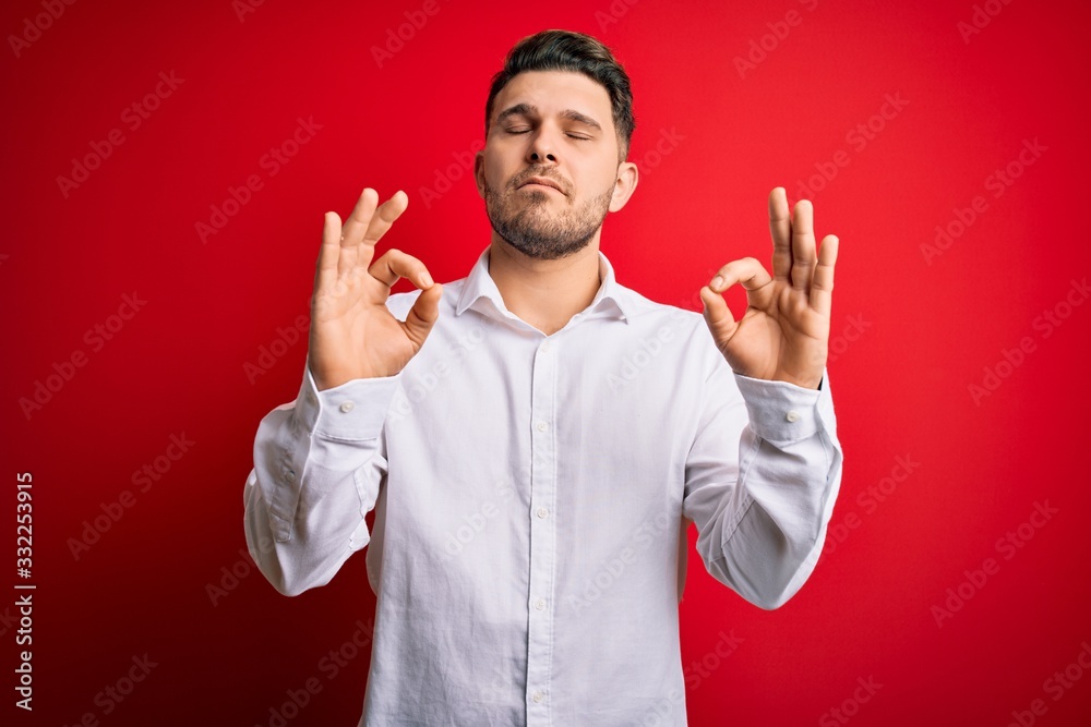 Young business man with blue eyes wearing elegant shirt standing over red isolated background relaxed and smiling with eyes closed doing meditation gesture with fingers. Yoga concept. Young business man with blue eyes wearing elegant shirt standing over red isolated background relaxed and smiling with eyes closed doing meditation gesture with fingers. Yoga concept.