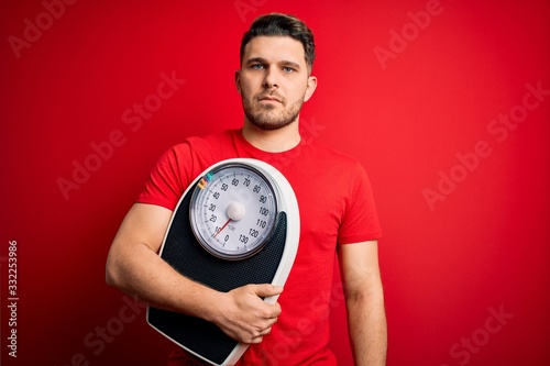 Young fitness man with blue eyes holding scale dieting for healthy weight over red background with a confident expression on smart face thinking serious