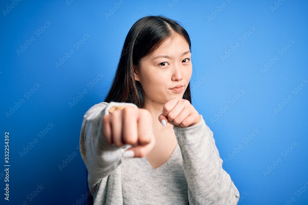 Fototapeta premium Young beautiful asian woman wearing casual sweater standing over blue isolated background Punching fist to fight, aggressive and angry attack, threat and violence