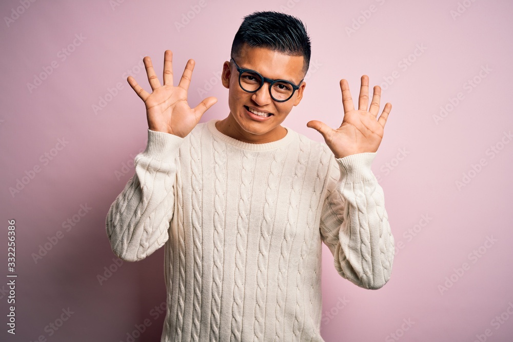 Naklejka premium Young handsome latin man wearing white casual sweater and glasses over pink background showing and pointing up with fingers number ten while smiling confident and happy.
