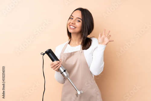 Young brunette girl using hand blender over isolated background saluting with hand with happy expression