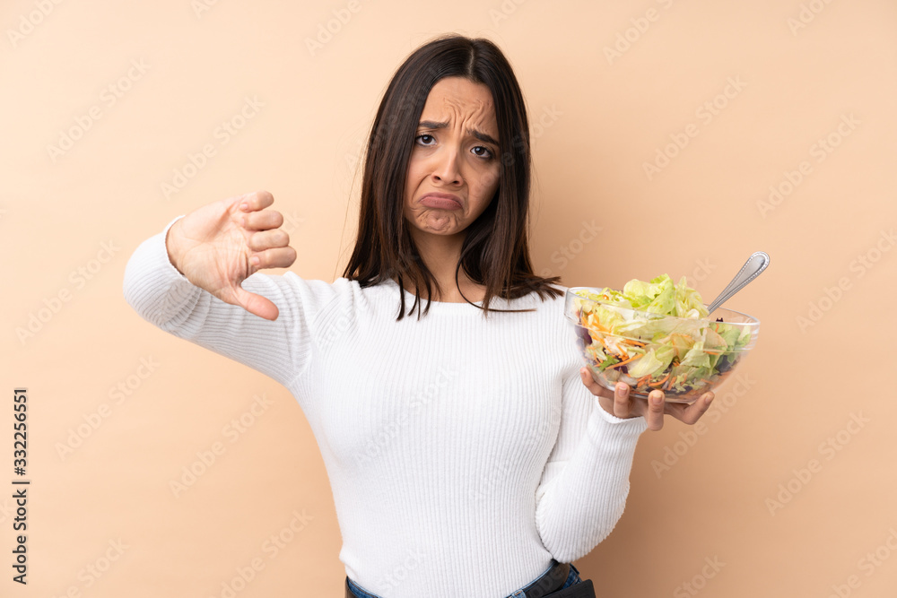 Young brunette girl holding a salad over isolated background showing thumb down Young brunette girl holding a salad over isolated background showing thumb down