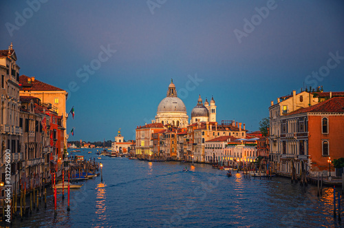 Venice cityscape with Grand Canal waterway. Buildings with evening lights along Grand Canal. Santa Maria della Salute Roman Catholic church on Punta della Dogana at twilight. Veneto Region, Italy.