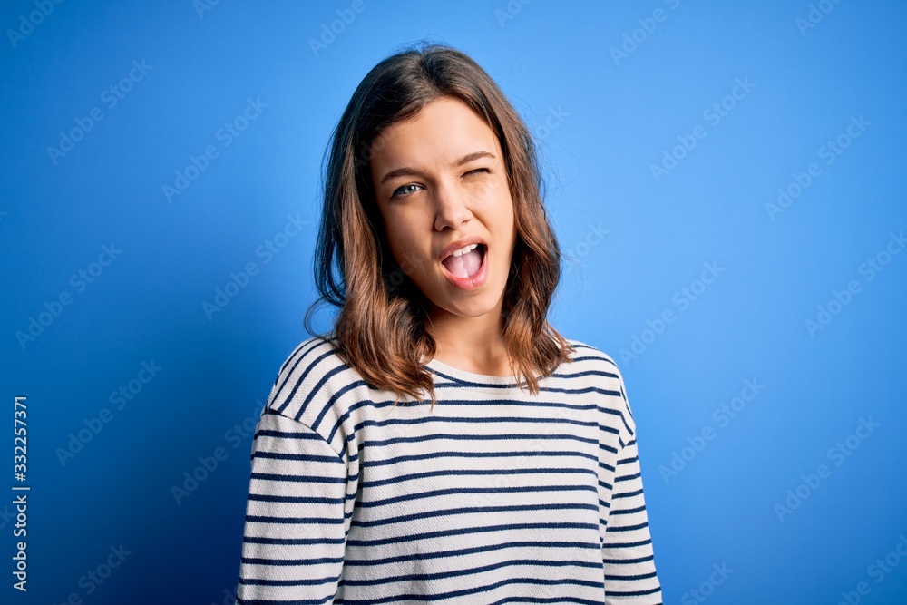 Young beautiful blonde girl wearing casual sweater standing over blue isolated background winking looking at the camera with sexy expression, cheerful and happy face. Young beautiful blonde girl wearing casual sweater standing over blue isolated background winking looking at the camera with sexy expression, cheerful and happy face.