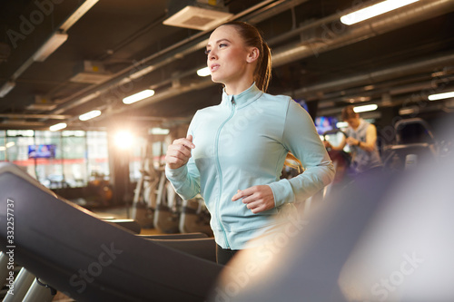 Portrait of sportive young woman running on treadmill during cardio workout in modern gym, lens flare