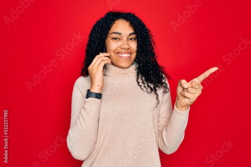 Young african american curly woman having conversation talking on the smartphone very happy pointing with hand and finger to the side