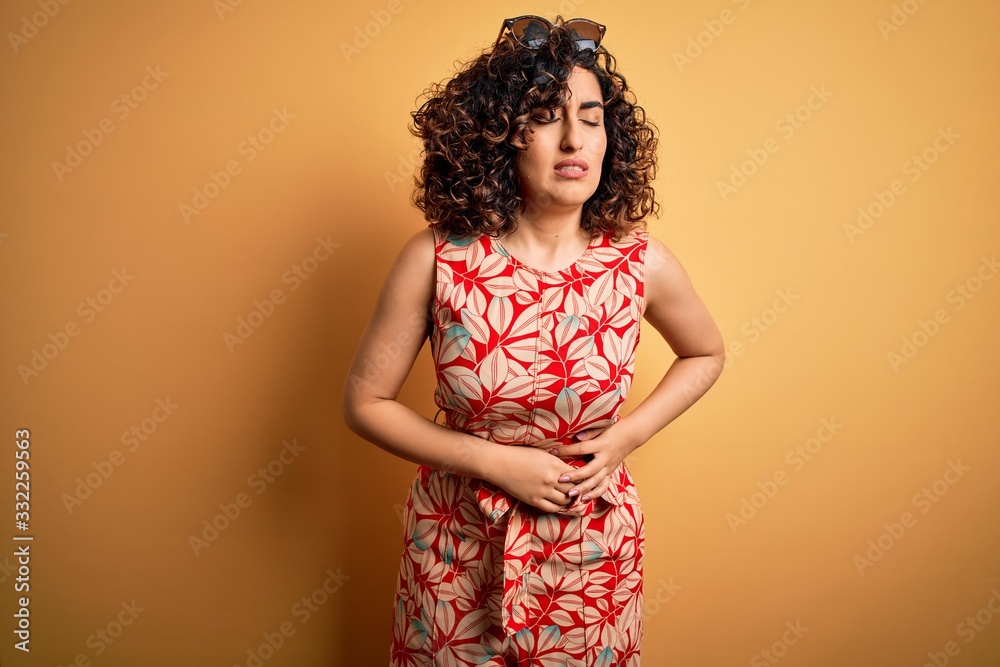 Young beautiful curly arab woman on vacation wearing summer floral dress and sunglasses with hand on stomach because indigestion, painful illness feeling unwell. Ache concept. Young beautiful curly arab woman on vacation wearing summer floral dress and sunglasses with hand on stomach because indigestion, painful illness feeling unwell. Ache concept.
