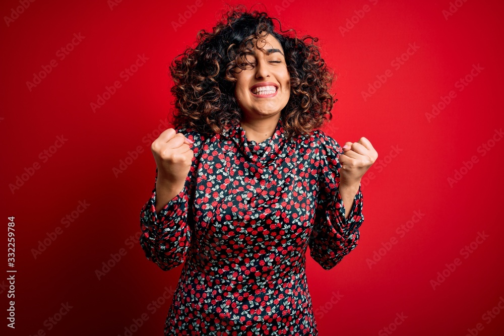 Young beautiful curly arab woman wearing casual floral dress standing over red background excited for success with arms raised and eyes closed celebrating victory smiling. Winner concept. Young beautiful curly arab woman wearing casual floral dress standing over red background excited for success with arms raised and eyes closed celebrating victory smiling. Winner concept.