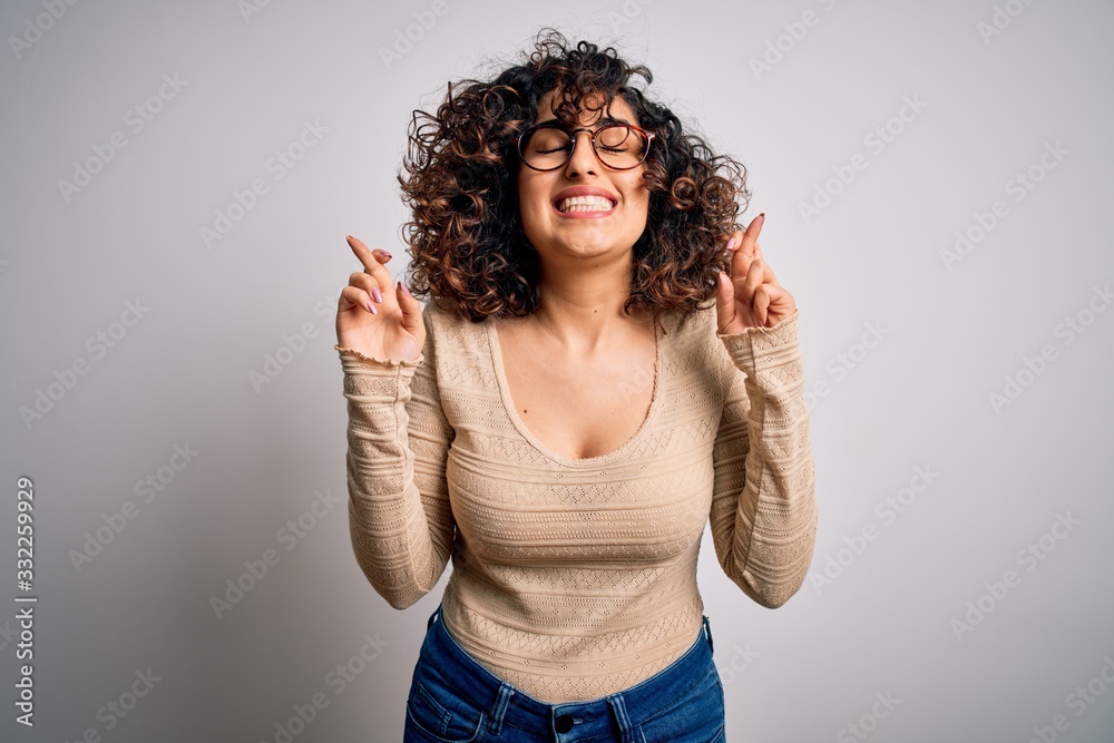 Young beautiful curly arab woman wearing casual t-shirt and glasses over white background gesturing finger crossed smiling with hope and eyes closed. Luck and superstitious concept.