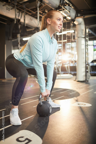 Full length portrait of smiling young woman picking up dumbbell during strength workout in sunlit gym, copy space